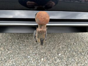 Rostige Anhängerkupplung eines schwarzen Autos, Nahaufnahme auf kiesigem Boden.