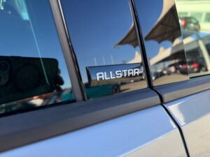 Nahaufnahme eines silbernen Autos mit "ALLSTAR"-Emblem. Reflektion einer Hand mit Kamera.